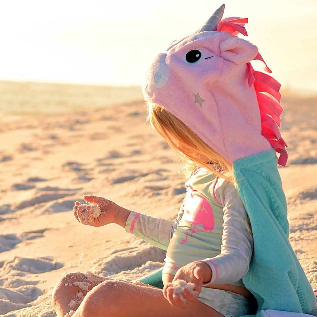 A child having a magical time playing in the sand at the beach while wearing the Zoocchini Alicorn hooded towel for kids